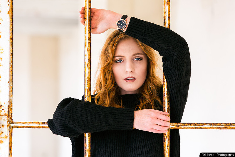 Young woman with red hair, looking at camera standing behind bars. Wearing a wrist watch on the inside of her wrist.