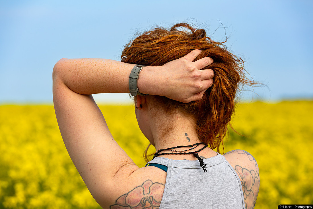 A woman with a semi-colon tattoo on her back stands in a field, symbolizing mental health awareness and resilience. Her watch is worn on the inside of her wrist as a daily reminder that change is possible.