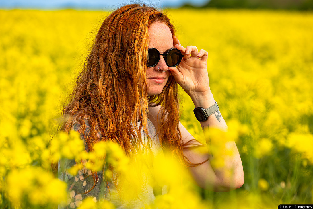 A woman with red hair and sunglasses stands in a field of yellow flowers, wearing her Fitbit inside her wrist as part of the Insider Watch Challenge.