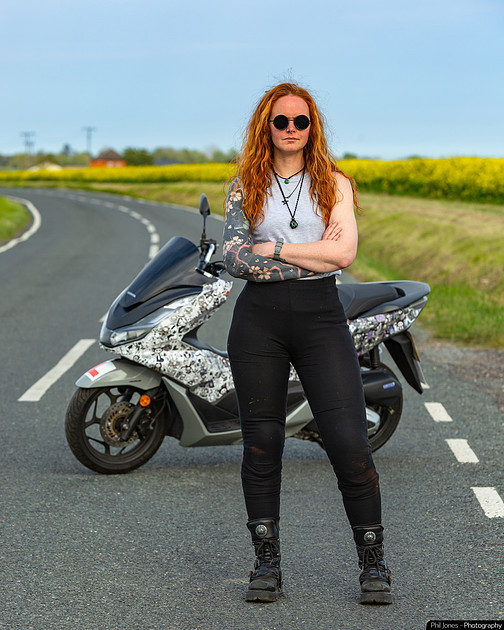 A woman stands beside a motorcycle, smiling and dressed in casual riding gear.