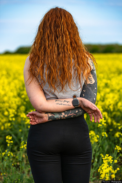 A woman with tattoos stands in a field of yellow flowers, showcasing her hypermobility in a vibrant, natural setting.