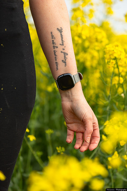 A woman with a wrist tattoo and a Fitbit worn inside the wrist watch stands in a vibrant field of yellow flowers, enjoying the sunny day.