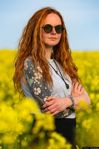 A woman with red hair and tattoos stands in a vibrant field of yellow flowers, enjoying the sunny day.