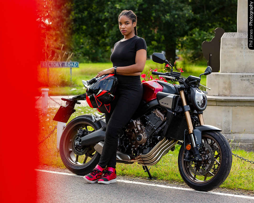 A woman leans against her red Honda motorcycle holding her protective helmet on a warm summers evening