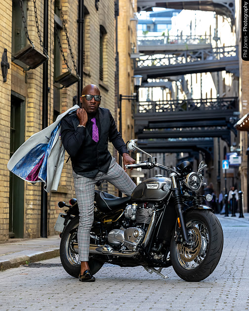 A man wearing smart attire steps off his motorcycle, holding a jacket over his shoulder, Triumph Speedmaster motorcycle