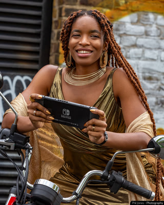 a woman sits on her motorcycle wearing a gold dress and holds a Nintendo Switch consul