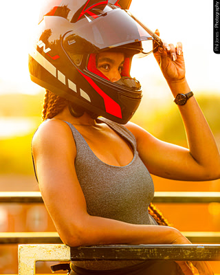 A woman wearing a motorcycle helmet leans on a barrier as the sun is setting, she wears a watch on the inside of her wrist