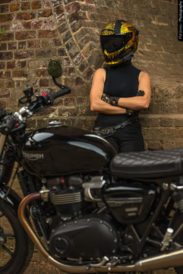 A female motorcyclist stands alongside a viaduct arch wearing her motorcycle clothing and crash helmet with bike in foreground