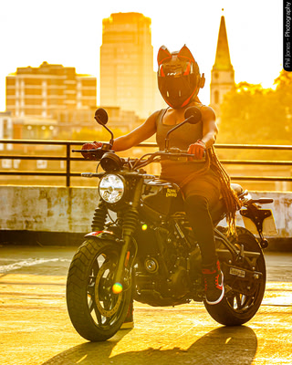 Person wearing a helmet with ears, riding a motorcycle in a parking lot at sunset, with city buildings in the background.
