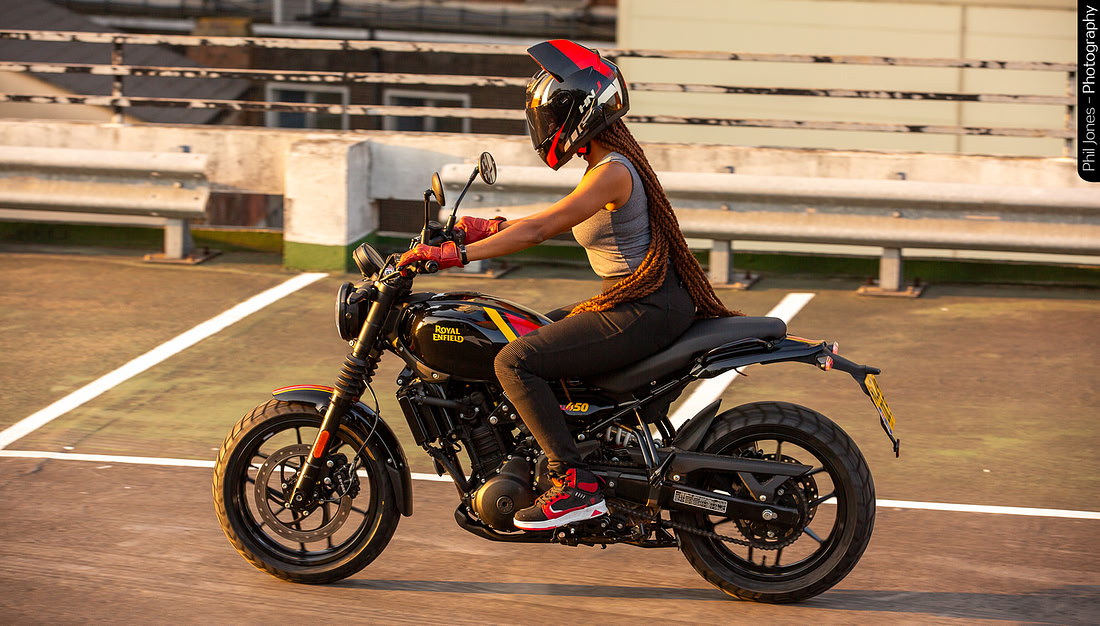 A person with long braided hair rides a black and red Royal Enfield motorcycle in a parking lot, wearing a helmet and casual clothing.