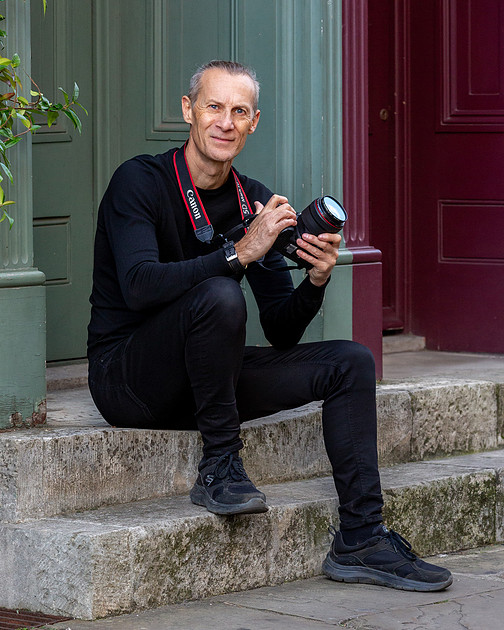 Photographer sitting on outdoor stone steps holding a Canon camera, wearing an all-black outfit with a red camera strap, in front of green and burgundy doors.