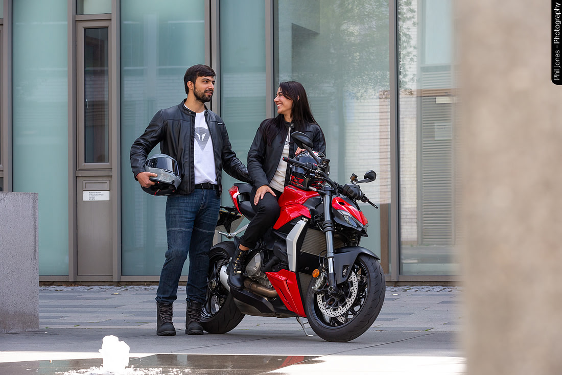 A man and a woman stand next to a motorcycle, both smiling and enjoying their time together in a vibrant setting.